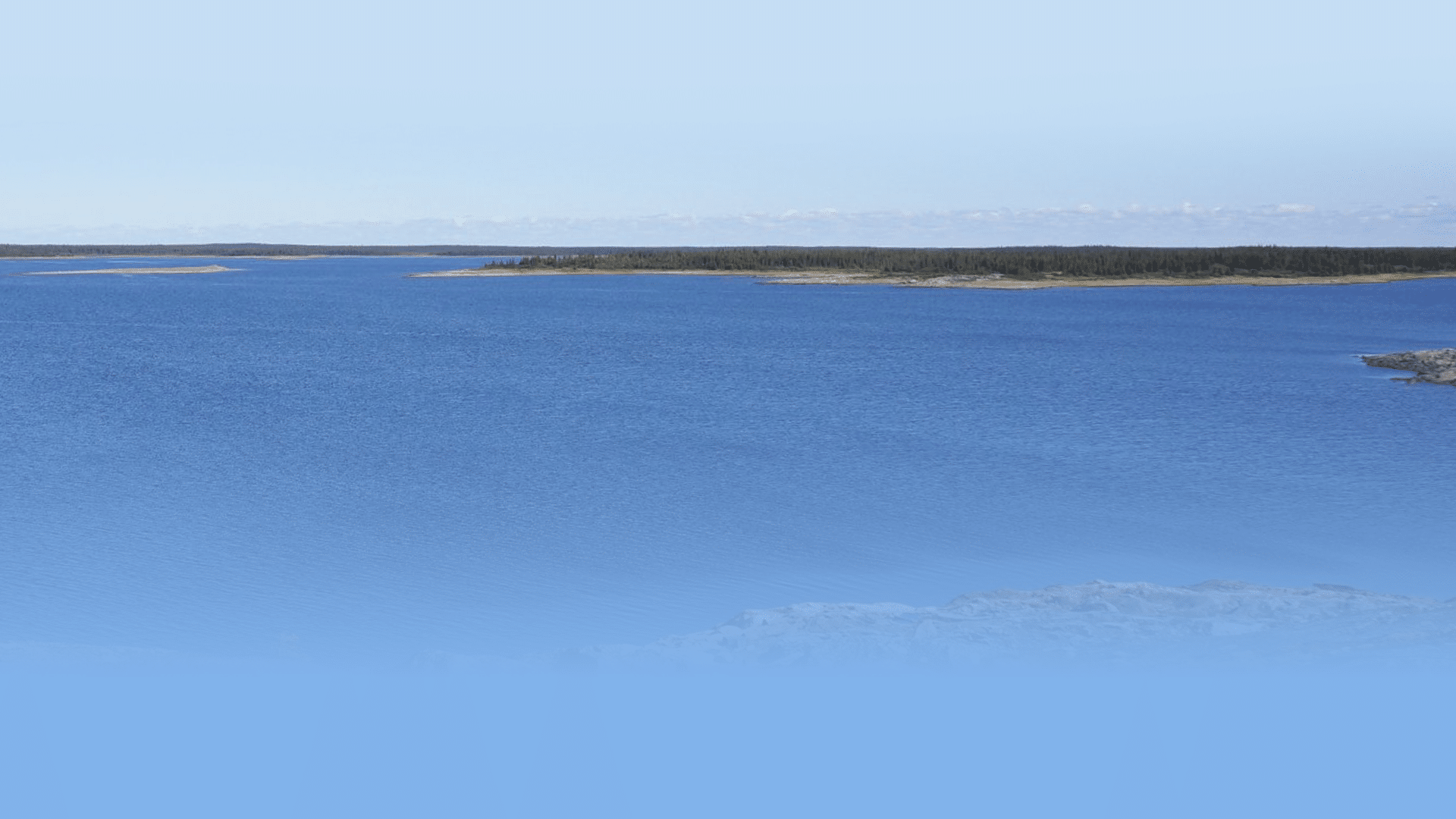 A serene view of a vast, calm blue lake under a clear sky. The lake stretches towards the horizon with distant, low-lying land covered in green vegetation. The foreground features rocky shores partially submerged in water.