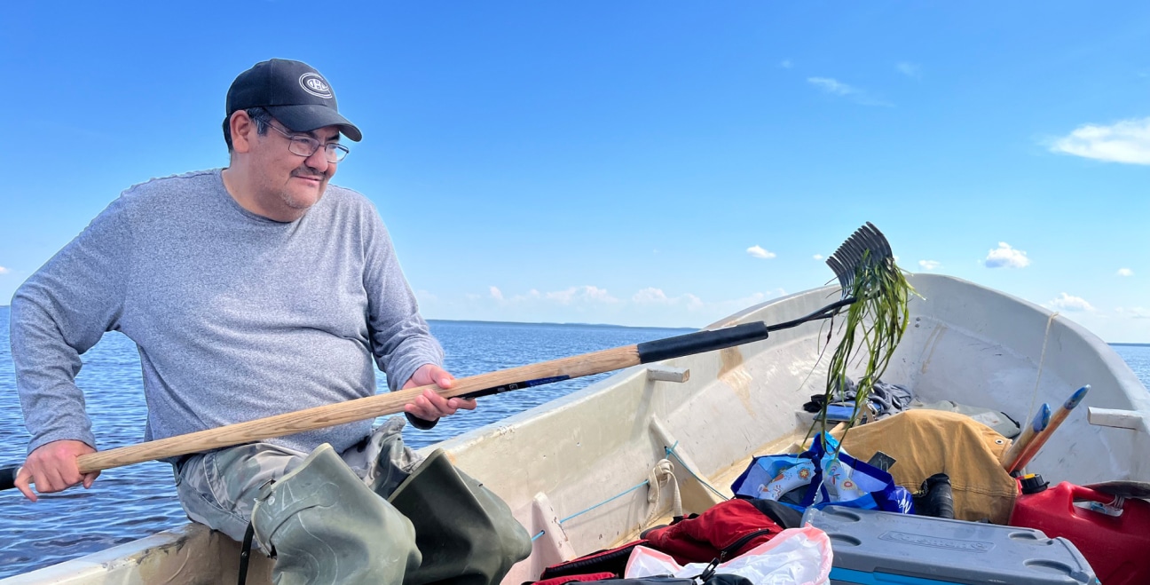 A man in a gray sweater and cap sits in a boat, holding a rake with aquatic plants. The boat is filled with supplies, and the water and blue sky are visible in the background.