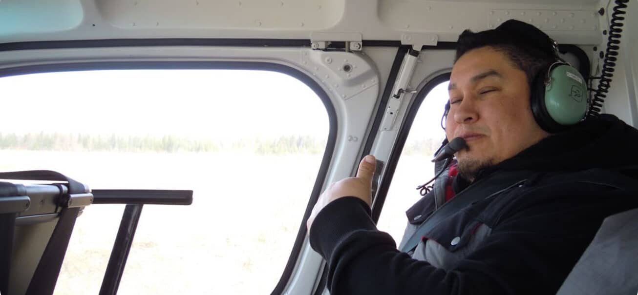 A person wearing headphones and a microphone is sitting inside a helicopter. They are giving a thumbs-up gesture and smiling. The background shows a window with a view of a light sky and blurry landscape.