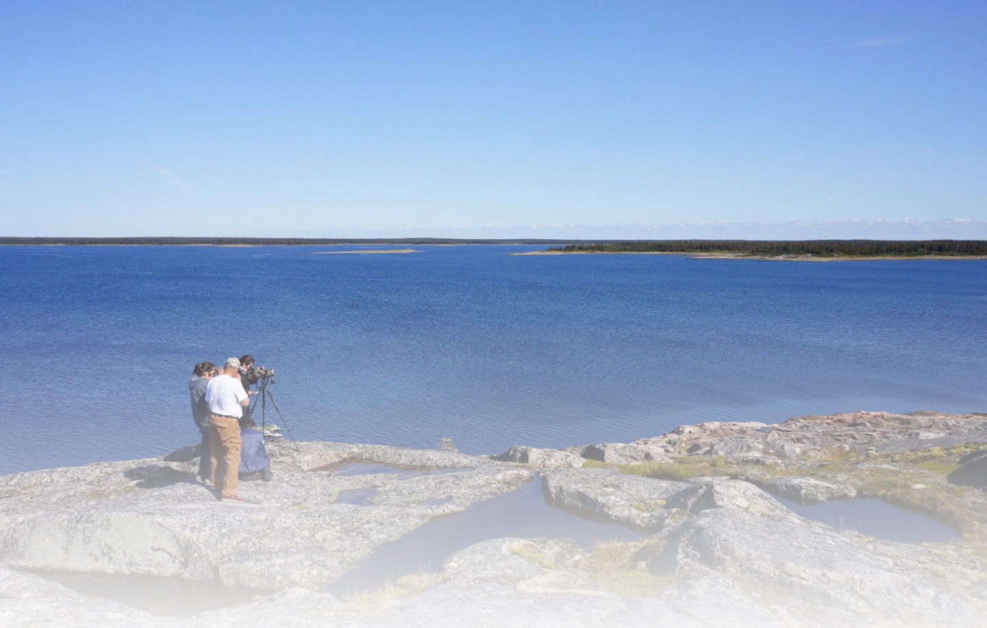 Two people with a tripod and camera equipment stand on rocky terrain overlooking a vast, calm blue lake under a clear blue sky. Distant shores and sparse trees line the horizon.