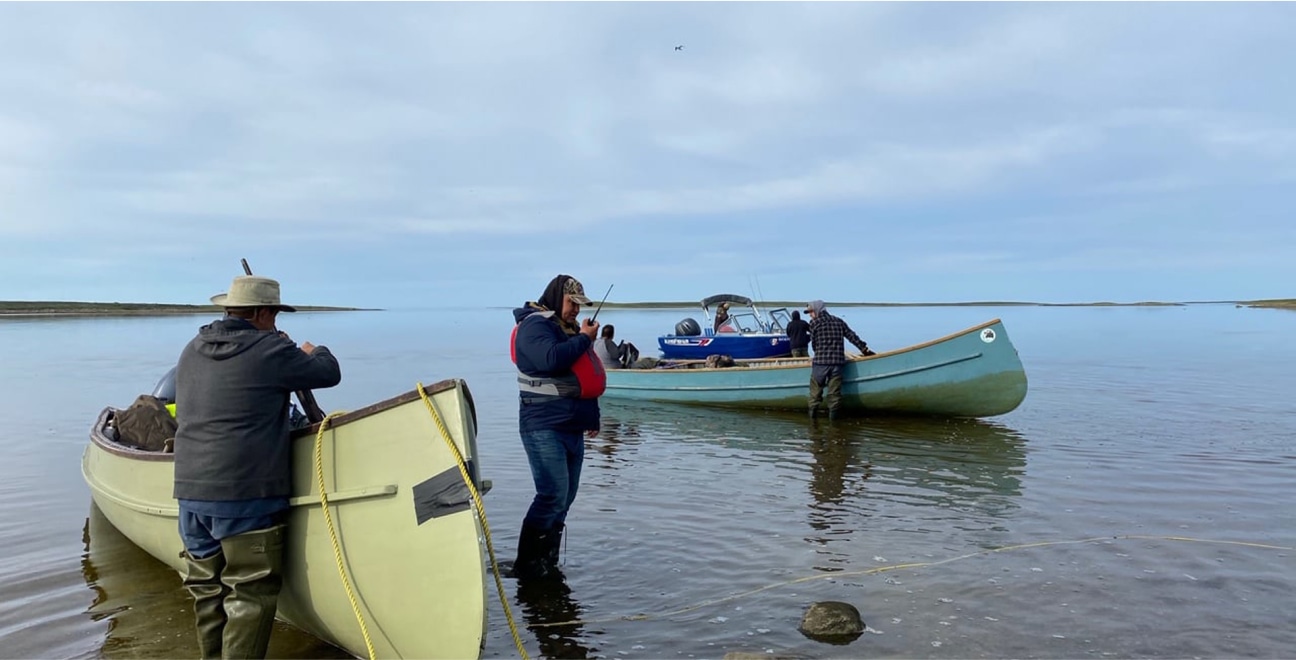 Three people stand in shallow water, maneuvering small boats near a calm shoreline under a cloudy sky. One person wears a life jacket, another holds a paddle, and the third is at the rear of a boat.